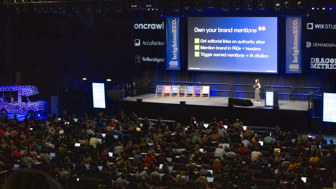 متحدّث على مسرح brightonSEO في لقطة من أعلى آخر قاعة ممتلئة، إلى جانب شريحة عرض ولافتتين طويلتين تحملان اسم brightonSEO.