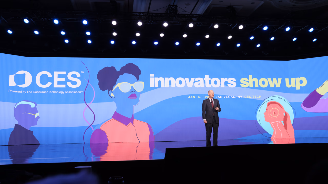 A speaker alone on a wide keynote stage at CES, in front of a blue illustrated screen reading «CES — innovators show up».