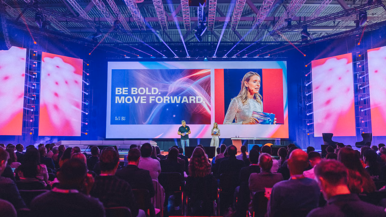 المسرح الرئيسي في DMEXCO من آخر قاعة ممتلئة: مقدّمان تحت شاشة عريضة كُتب عليها «Be bold. Move forward.»، والقاعة مضاءة بالأرجواني والأزرق.