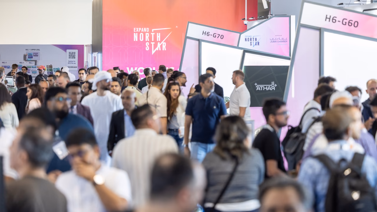 A dense crowd of visitors between white startup stands at Expand North Star, beneath a pink and orange pylon carrying the event's logo.