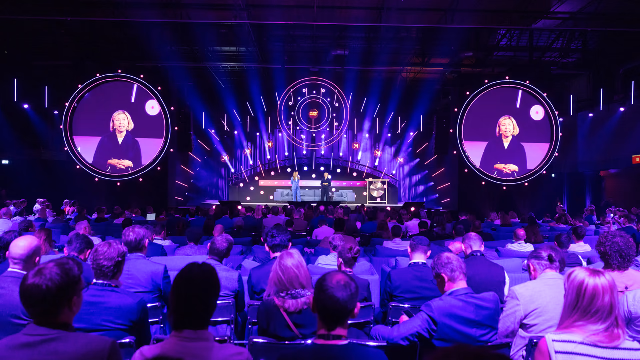 المسرح الرئيسي في Money20/20 Europe من آخر قاعة ممتلئة بإضاءة بنفسجية ووردية، ويظهر أحد المتحدّثين على شاشتين دائريتين كبيرتين.