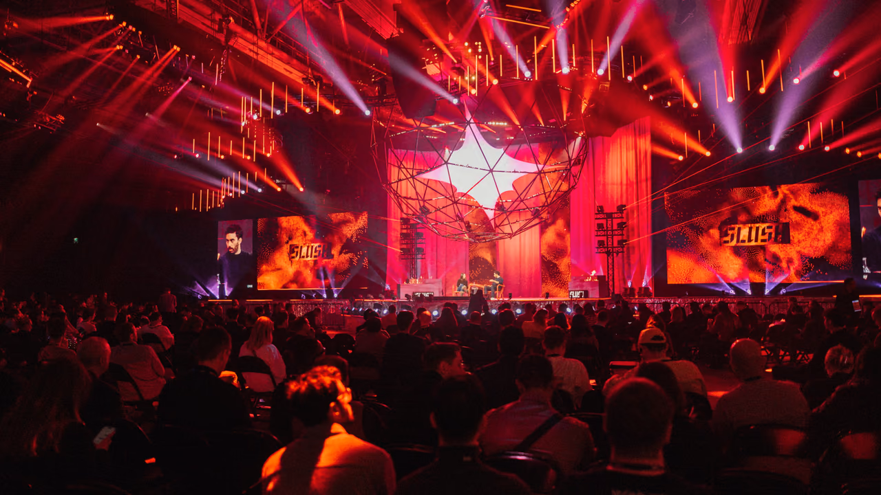 Slush's Founder Stage seen from the back of a packed, darkened hall: the room washed deep red by an overhead lighting rig, a glowing star-shaped canopy above the stage, and SLUSH on the screens either side.