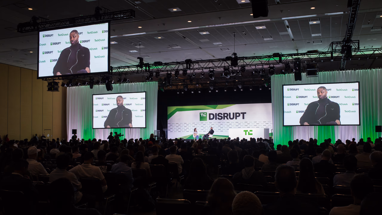 المسرح الرئيسي في TechCrunch Disrupt من آخر قاعة ممتلئة في Moscone West: خلفية Disrupt مضاءة بالأخضر وثلاث شاشات فوق جمهور جالس.