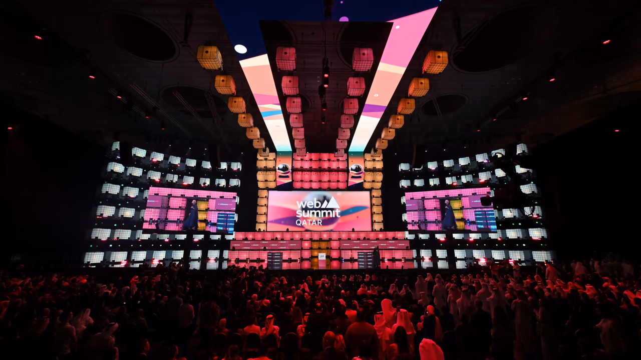 Web Summit Qatar's Centre Stage in Doha: a pink and orange set of stacked cubes with «web summit Qatar» on its central screen, above a standing crowd.