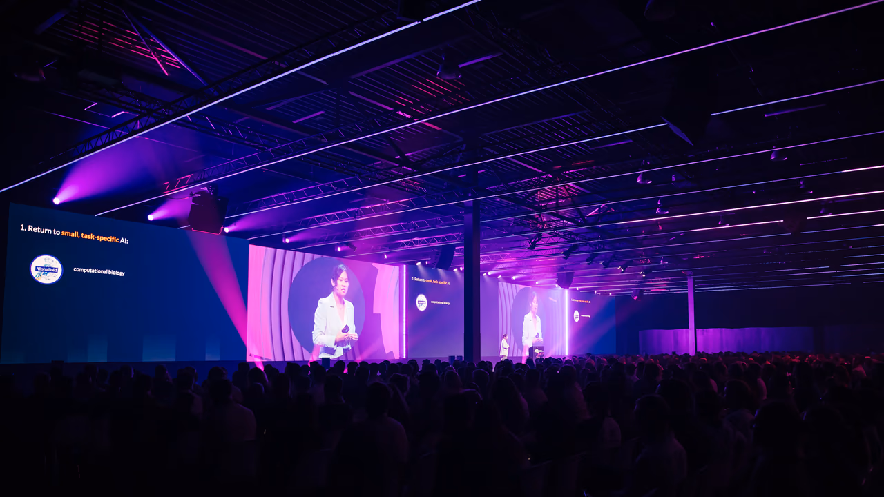 World Summit AI's main stage from the back of a dark, full hall: a speaker on a long run of screens, washed in purple light.