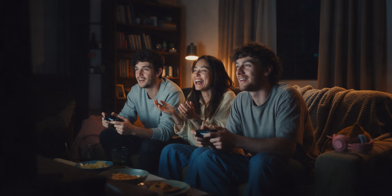 ثلاثة أصدقاء على أريكة في غرفة معيشة خافتة الإضاءة، اثنان يمسكان بذراعي تحكم وثالثة تضحك، ووجوههم مضاءة بشاشة خارج الإطار.