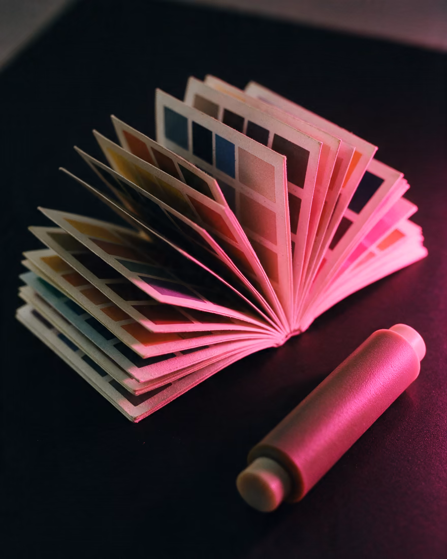 Colour swatch books fanned open beside an ink roller on black, raked by pink light.