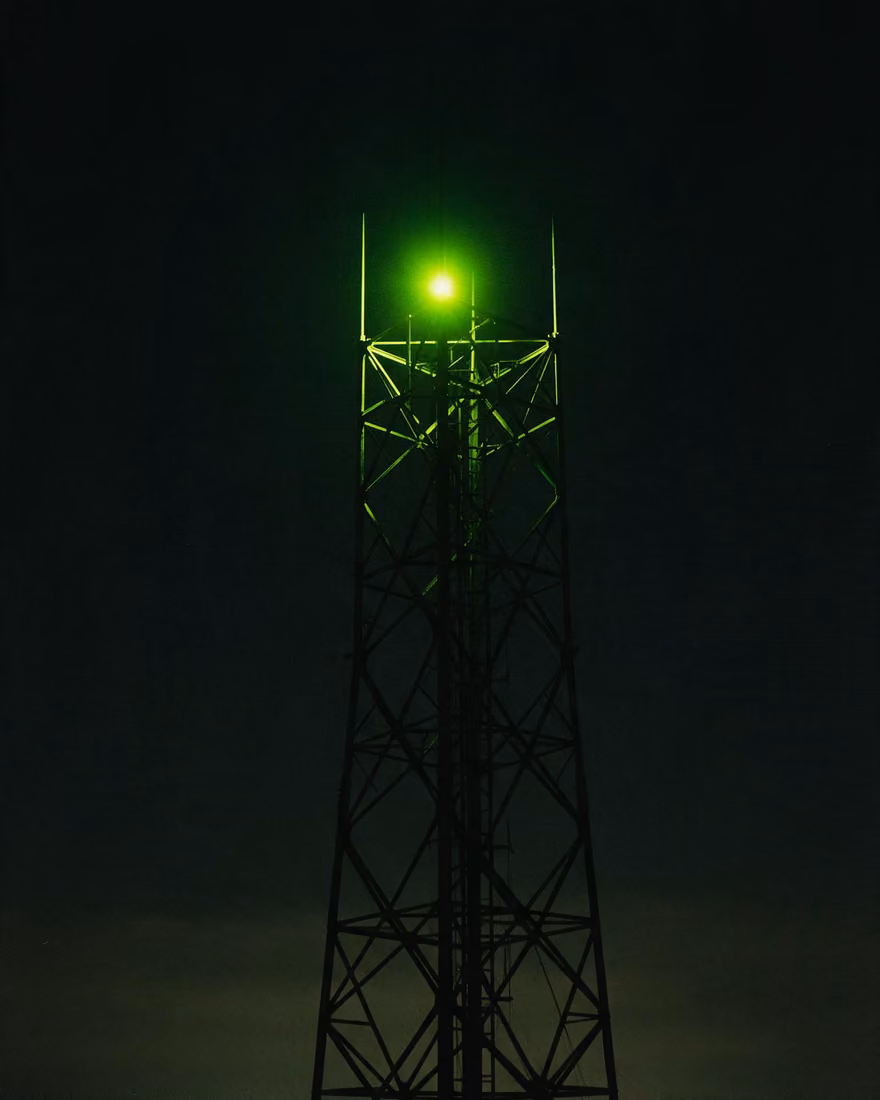 A steel communications mast at night, black against the sky, one green beacon burning near the top.