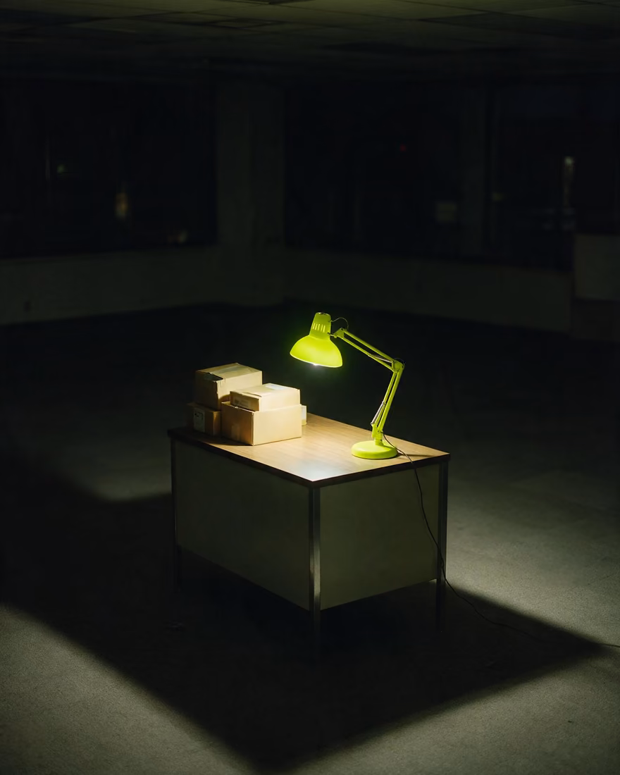 One desk and a few unopened boxes under a green work lamp on an otherwise empty floor.